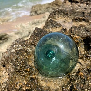 Könnte beinhalten: Eine kugelförmige, durchscheinende Fischerei-Schwimmkugel aus meergrünem Glas mit einer deutlichen kreisförmigen Siegelmarkierung auf der Oberfläche. Das Objekt ist glatt und abgerundet, reflektiert das Licht und zeigt einen tiefen Farbton.