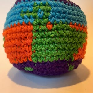 May include: A spherical crochet ball featuring a multi-colored geometric pattern. The yarn colors include bright orange, vibrant green, sky blue, and deep purple, worked in a dense, textured stitch that creates a firm, rounded shape.