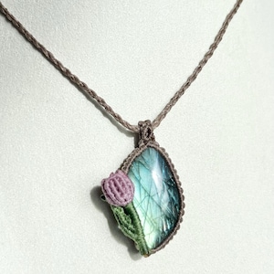 May include: A handcrafted macrame necklace featuring a teardrop-shaped labradorite pendant with blue and green iridescence. A small, crocheted purple flower with green stem accents the pendant. The necklace cord is a neutral brown color.