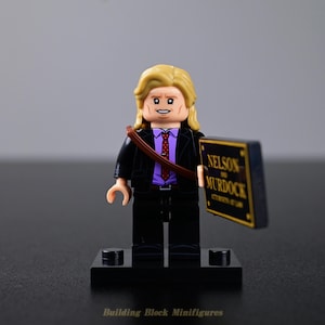 Op de afbeelding: Een plastic minifiguur met blond haar, gekleed in een zwart colbert over een paars overhemd met een rood gestreepte das. Het figuurtje houdt een rechthoekig zwart bordje vast met de gouden tekst NELSON AND MURDOCK ATTORNEYS AT LAW.