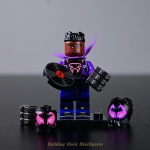 Peut inclure: Une figurine de bloc de construction en plastique avec une peau foncée, des cheveux noirs et une cape violette. Le personnage porte une tenue noire et violette avec un emblème rose, un pantalon bleu et tient un disque vinyle noir.