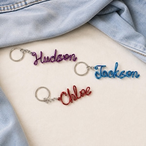 May include: Three metal wire keychains featuring the names Hudson in purple, Jackson in blue, and Chloe in red. Each keychain consists of a silver-toned split ring attached to a continuous piece of colored wire bent into cursive script lettering.
