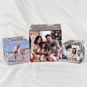 Op de afbeelding: Drie aangepaste fotoblokken met afgedrukte familiefoto's. Het linkerblok toont een persoon en kind op een strand. Het middelste blok toont een gezin in pyjama bij een kerstboom. Het rechterblok toont twee kinderen die met blokken spelen.