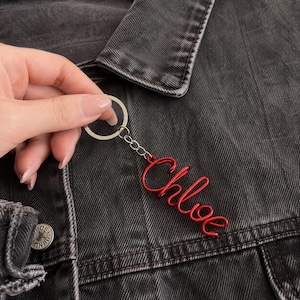 Puede incluir: Un llavero de alambre rojo con el nombre Chloe en letra cursiva. El llavero está unido a una anilla de metal plateado mediante una cadena corta. El diseño está formado por una sola pieza continua de alambre de color rojo.