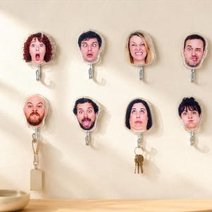 May include: A set of eight wall-mounted hooks, each featuring a clear acrylic plaque printed with a unique human face displaying a different facial expression. The hooks are arranged in two rows, with some holding metal keys or a blank tag.