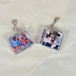 Könnte beinhalten: Zwei quadratische Foto-Schlüsselanhänger aus klarem Acryl mit silbernen Kugelkettchen. Der linke Anhänger zeigt eine vierköpfige Familie am Strand. Der rechte Anhänger zeigt eine vierköpfige Familie, die gemeinsam in einem Innenraum sitzt.