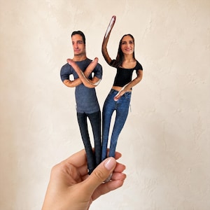 May include: Two custom fabric dolls featuring printed photographic portraits of a person with dark hair wearing a grey t-shirt and black pants, and a person with long dark hair wearing a black top and blue jeans, both with arms raised.