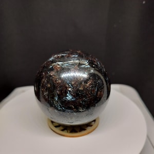 May include: A polished spherical stone with a dark, mottled surface featuring deep brown and black tones. The sphere displays a reflective, metallic sheen with visible internal crystalline structures and light-catching streaks across its rounded form.