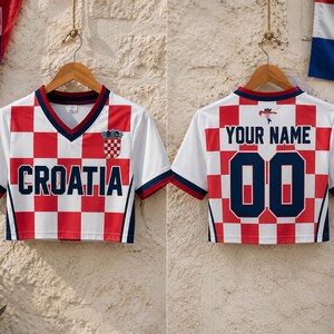 Puede incluir: Un top corto de manga corta con cuello en V y estampado de cuadros rojos y blancos. La parte delantera muestra el texto CROATIA en azul marino con un peque&ntilde;o escudo. La parte trasera muestra el texto YOUR NAME sobre el n&uacute;mero 00.