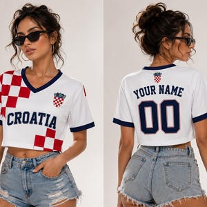 Puede incluir: Una camiseta corta blanca con cuello en V, con patrones de cuadros rojos y blancos en la parte delantera, el texto CROATIA en azul marino y un peque&ntilde;o escudo. La parte trasera muestra el texto YOUR NAME sobre el n&uacute;mero 00 en azul marino.