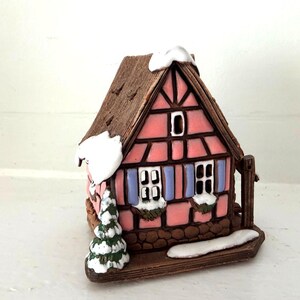 Può includere: Una statuetta di casa in miniatura in ceramica con facciata rosa e intelaiatura in legno marrone, tetto marrone testurizzato coperto di neve bianca, persiane azzurre, due finestre e un piccolo pino verde attaccato al lato della struttura.