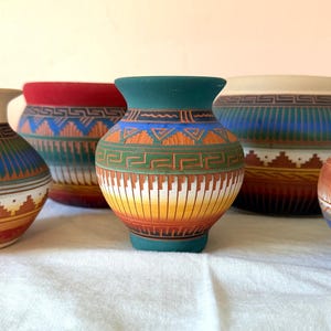 May include: A collection of five small ceramic vases featuring matte finishes and intricate geometric patterns. The vessels display vibrant color gradients including teal, red, orange, blue, and cream, with etched Greek key and sawtooth motifs.