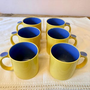 May include: A set of six ceramic mugs arranged in two rows of three. Each mug features a bright yellow exterior and a contrasting blue interior. The handles are yellow and topped with a small, flat, circular blue accent piece at the top.