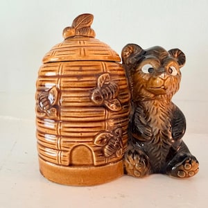 May include: A brown ceramic honey jar shaped like a beehive with a lid featuring a bee topper. The jar is decorated with embossed bees on the exterior and is attached to a brown ceramic bear figure sitting upright next to the hive structure.
