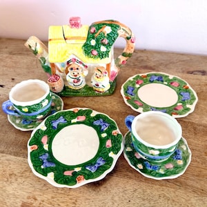May include: A ceramic tea set featuring a teapot shaped like a cottage with two bear figures, two cups, and two saucers. The pieces are decorated with green vines, pink flowers, and blue bows on a white background with yellow roof details.