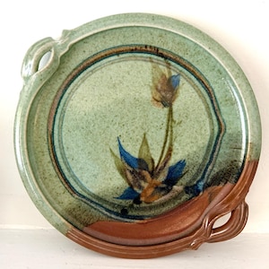 Può includere: Un piatto da portata rotondo in ceramica con smalto verde chiaro maculato e un motivo floreale dipinto a mano in blu e marrone al centro. Il piatto presenta due manici integrati sui lati opposti e una base in smalto marrone a contrasto.