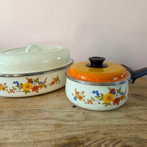 May include: An oval cream-coloured enamel roasting pan with a matching lid and a round cream-coloured enamel saucepan with a bright orange lid and black handle. Both pieces feature a colourful floral pattern of yellow, orange, and blue flowers.