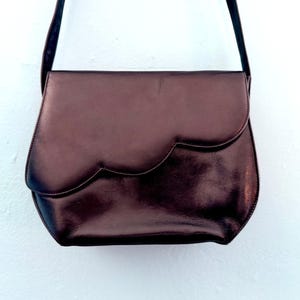 May include: A dark metallic bronze shoulder bag with a smooth, polished finish. The bag features a distinctive scalloped flap closure that curves across the front, a rounded bottom silhouette, and a thin, matching shoulder strap attached at the top.