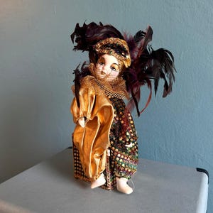 Op de afbeelding: Een porseleinen clownspop met een beschilderd gezicht met traanmarkeringen, gekleed in een goudkleurig satijnen en paillettenkostuum met een ruchekraag. De pop is versierd met een hoofdtooi met donkerpaarse en zwarte veren.