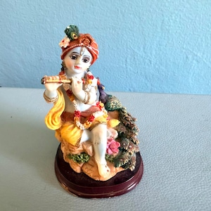May include: A figurine of a deity playing a flute, wearing an orange turban with a peacock feather, a yellow garment, and a floral garland. The figure sits on a rock base next to a peacock with detailed feathers, all mounted on a dark circular stand.