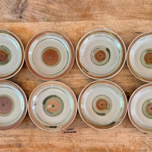 May include: A set of eight round ceramic plates arranged in two rows. Each plate features a light gray base with a prominent reddish-brown circular center, surrounded by a subtle green ring and a reddish-brown rim along the outer edge.