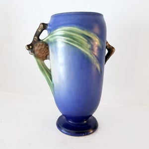 May include: A blue ceramic vase with a matte finish, featuring a raised relief design of a brown pinecone and green pine needles on the side. The vase has a pedestal base and a smooth, rounded body, showcasing a blend of blue, green, and brown tones.