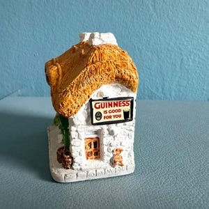 Pode incluir: Uma estatueta em miniatura de uma casa de pedra branca com um telhado de palha amarelo texturizado. Uma placa retangular na parede frontal apresenta o texto preto e vermelho GUINNESS IS GOOD FOR YOU com um copo de cerveja e uma porta castanha.