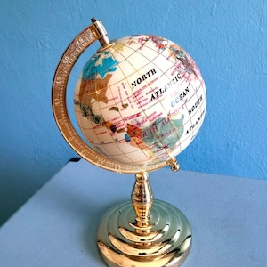 Op de afbeelding: Een tafelglobe met een crèmekleurige bol met veelkleurige continenten en oceanen, gemonteerd op een glanzende goudkleurige metalen standaard. De gebogen meridiaanboog bevat gegraveerde graadmarkeringen en de voet is een ronde, getrapte metalen voet.