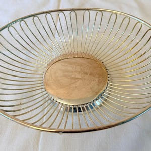 Può includere: Un cestino rotondo in filo metallico con una finitura dorata lucida. Il design è costituito da una solida piastra di base circolare circondata da sottili raggi verticali in filo metallico, distanziati uniformemente, che si curvano verso l'alto.