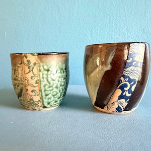 May include: Two ceramic cups with distinct finishes. The left cup features a textured green glaze with a vertical band of light-colored floral patterns. The right cup displays a smooth dark brown glaze contrasted by a vertical panel of blue and white floral motifs.
