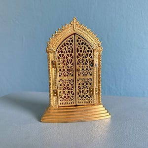 Puede incluir: Un marco de metal dorado estilo tríptico con puertas de filigrana calada en forma de corazón. La estructura tiene una parte superior en arco apuntado, una base escalonada y bisagras laterales visibles que revelan un interior de color rojo.