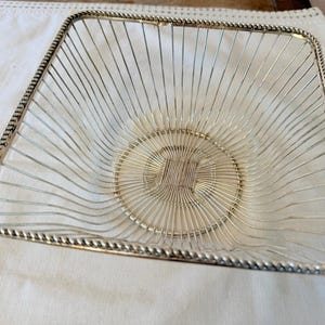 May include: A square silver-toned wire basket featuring a decorative twisted rope-style rim. The basket is constructed from thin, vertical metal wires that converge into a circular, woven base pattern, creating an open, airy, and geometric design.
