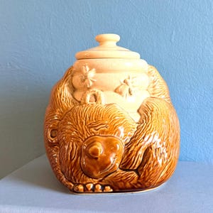 Può includere: Un barattolo per miele in ceramica raffigurante un orso marrone lucido raggomitolato davanti a un alveare color crema. L'alveare presenta una superficie testurizzata con api scolpite e un coperchio abbinato con un piccolo pomello. Finitura smaltata.