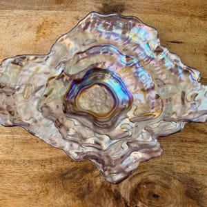 May include: An iridescent glass bowl with a wavy, organic edge and a rippled surface texture. The glass features a multi-colored sheen with purple, blue, and gold tones. A small circular sticker is visible on the left side of the bowl's rim.