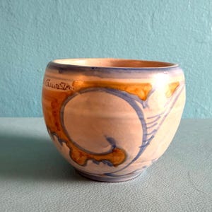 May include: A rounded ceramic bowl with a cream-colored base featuring hand-painted swirling patterns in shades of blue and golden yellow. The exterior surface displays a visible signature near the rim, with a smooth, curved silhouette.