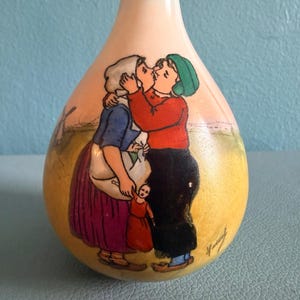 May include: A pear-shaped ceramic vase featuring a hand-painted scene of two figures kissing in a field with a windmill in the background. The figures wear traditional clothing, including a white head covering, a blue top, and a red sweater.