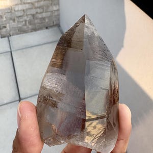 Puede incluir: Una gran punta de cristal de cuarzo ahumado semitransparente con un ápice puntiagudo y facetas geométricas naturales. El mineral muestra tonos variados de marrón y gris translúcido, con inclusiones internas y bordes claros y afilados.