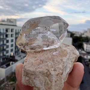 Puede incluir: Un esp&eacute;cimen de cristal de cuarzo transparente y facetado unido a una matriz mineral rugosa de color tostado. El cristal presenta inclusiones internas visibles y bordes geom&eacute;tricos afilados, contrastando con la base terrosa y texturizada.