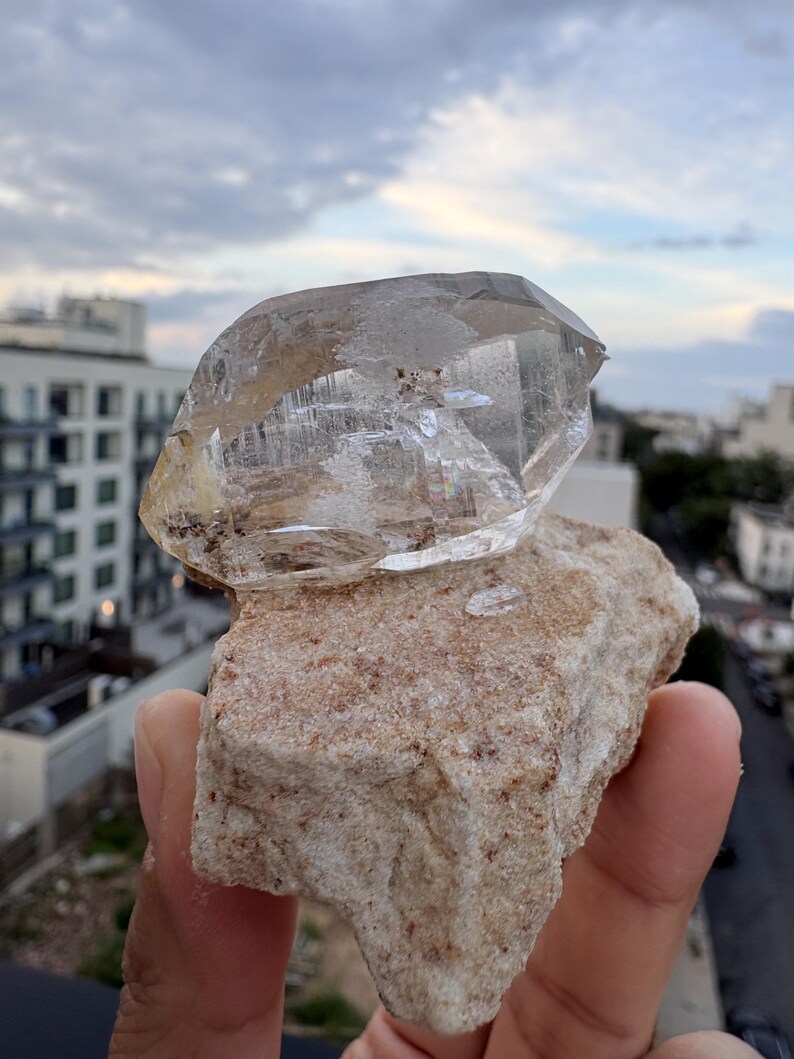 Puede incluir: Un esp&eacute;cimen de cristal de cuarzo transparente y facetado unido a una matriz mineral rugosa de color tostado. El cristal presenta inclusiones internas visibles y bordes geom&eacute;tricos afilados, contrastando con la base terrosa y texturizada.