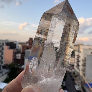 Puede incluir: Una punta de cristal de cuarzo hexagonal transparente con fracturas internas visibles, estrías horizontales a lo largo de los lados y una terminación puntiaguda. El cristal muestra transparencia natural con sutiles inclusiones marrones.