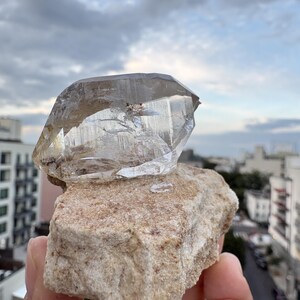 Puede incluir: Un esp&eacute;cimen de cristal de cuarzo transparente y multifac&eacute;tico unido a una matriz mineral rugosa de color tostado. El cristal presenta fracturas e inclusiones internas visibles, con una textura v&iacute;trea y transparente que contrasta con la base de piedra.