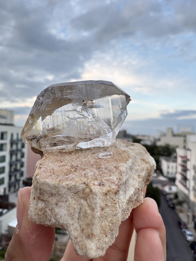 Puede incluir: Un esp&eacute;cimen de cristal de cuarzo transparente y multifac&eacute;tico unido a una matriz mineral rugosa de color tostado. El cristal presenta fracturas e inclusiones internas visibles, con una textura v&iacute;trea y transparente que contrasta con la base de piedra.
