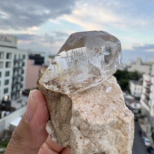Puede incluir: Una punta de cristal de cuarzo transparente y multifac&eacute;tica unida a una matriz de roca rugosa de color beige. El cristal presenta inclusiones internas visibles y bordes geom&eacute;tricos afilados, contrastando con la base terrosa y texturizada de la piedra.