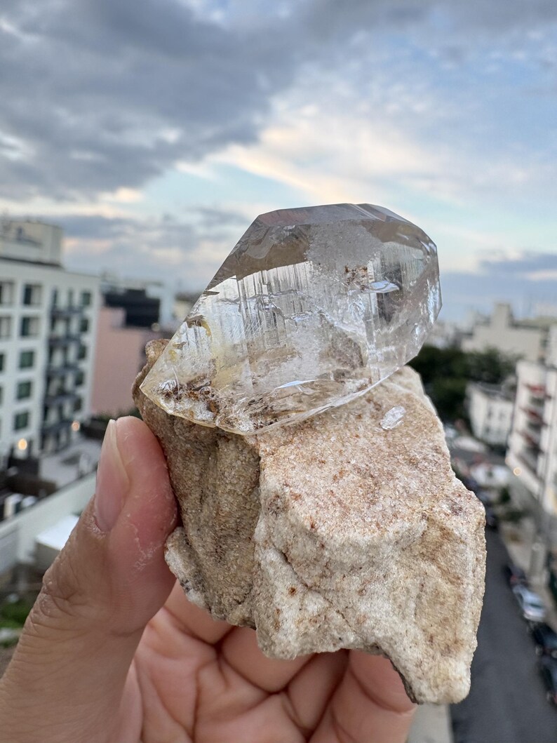 Puede incluir: Una punta de cristal de cuarzo transparente y multifac&eacute;tica unida a una matriz de roca rugosa de color beige. El cristal presenta inclusiones internas visibles y bordes geom&eacute;tricos afilados, contrastando con la base terrosa y texturizada de la piedra.