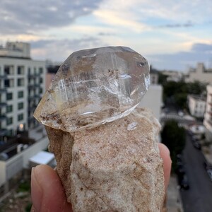 Puede incluir: Una punta de cristal de cuarzo transparente y facetada unida a una matriz mineral rugosa de color tostado. El cristal muestra fracturas internas e inclusiones naturales, mientras que la base tiene una textura arenosa y terrosa.
