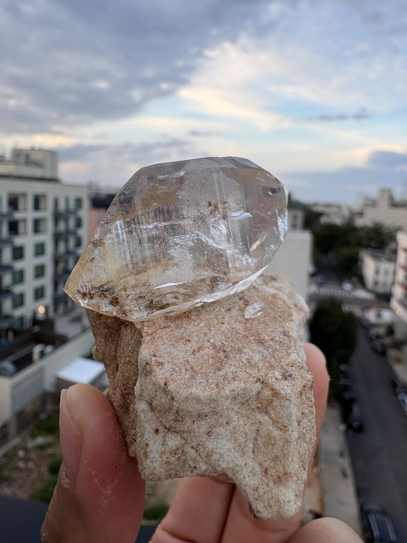 Puede incluir: Una punta de cristal de cuarzo transparente y facetada unida a una matriz mineral rugosa de color tostado. El cristal muestra fracturas internas e inclusiones naturales, mientras que la base tiene una textura arenosa y terrosa.