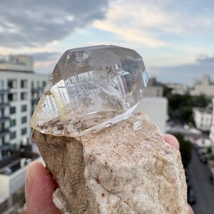 Puede incluir: Una punta de cristal de cuarzo transparente y facetado unida a una matriz mineral rugosa de color beige. El cristal presenta inclusiones internas visibles y bordes geom&eacute;tricos afilados, mientras que la base tiene una textura terrosa.