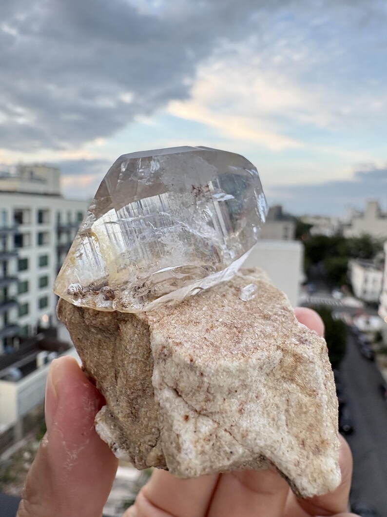 Puede incluir: Una punta de cristal de cuarzo transparente y facetado unida a una matriz mineral rugosa de color beige. El cristal presenta inclusiones internas visibles y bordes geom&eacute;tricos afilados, mientras que la base tiene una textura terrosa.