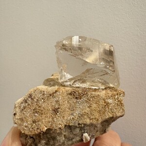 Puede incluir: Un esp&eacute;cimen de cristal angular, claro y transl&uacute;cido, unido a una matriz mineral rugosa, texturizada y de color tostado. El cristal presenta facetas geom&eacute;tricas distintas y claridad interna, contrastando con la roca base, gruesa y terrosa.