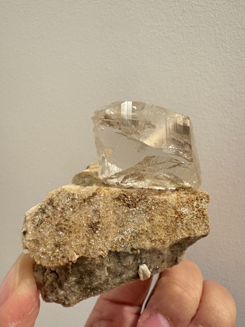 Puede incluir: Un esp&eacute;cimen de cristal angular, claro y transl&uacute;cido, unido a una matriz mineral rugosa, texturizada y de color tostado. El cristal presenta facetas geom&eacute;tricas distintas y claridad interna, contrastando con la roca base, gruesa y terrosa.